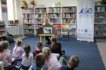 Zdjęcie przedstawia grupę dzieci i osobę dorosłą w Bibliotece. Kobieta siedzi za drewnianym teatrzykiem i opowiada