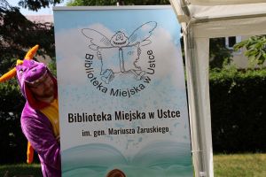 Baner z logiem i nazwą Biblioteka Miejska w Ustce im. gen. Mariusza Zaruskiego, za niego wyłania się bibliotekarz przebrany za smoka
