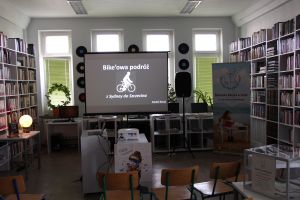 Zdjęcie przedstawia wnętrze Czytelni dla Dorosłych: ekran, regały z książkami i rozstawione krzesła. Na ekranie wyświetlony pierwszy  slajd prezentacji.