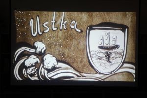 Zdjęcie na którym widzimy herb Ustki, fale oraz napis Ustka wszystko usypane z piasku 