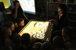 Zdjęcie na którym widzimy przyglądające się z bliska stołowi na którym tworzy się kolejne obrazy historii, malując je na piasku i za pomocą piasku