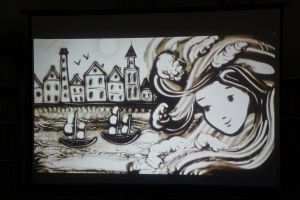 Zdjęcie na którym widzimy starą rybacką osadę, twarz Bryzgi Rosowej oraz statki w porcie wszystko usypane z piasku