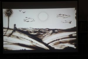 Zdjęcie na którym widzimy plażę, morze, ptaki oraz kobietę wpratrującą się w morze wszystko usypane z piasku