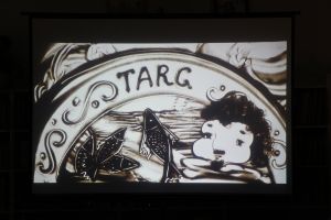 Zdjęcie na którym widzimy twarz starej kobiety, łososie oraz napis targ na okrągłym szyldzie wszystko usypane z piasku 