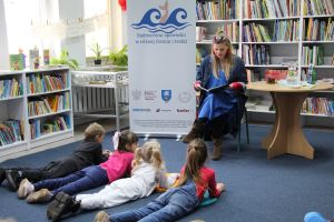 Zdjęcie przedstawia grupę dzieci i osobę dorosłą w Bibliotece. Kobieta czyta dzieciom opowieść.