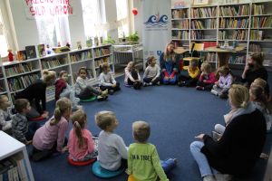 Zdjęcie przedstawia grupę dzieci i osoby dorosłe w Bibliotece. Wszyscy siedzą w kole na podłodze i rozmawiają.