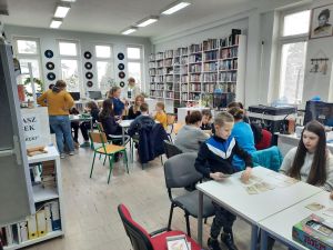 Zdjęcie przedstawia osoby grające w gry planszowe podczas cotygodniowych spotkań integracyjnych w Bibliotece.